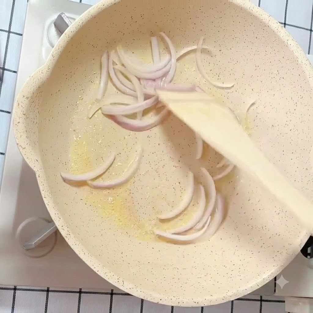 木製のスパチュラを使い、バターを引いたフライパンで白と紫の玉ねぎのスライスを炒めている様子。