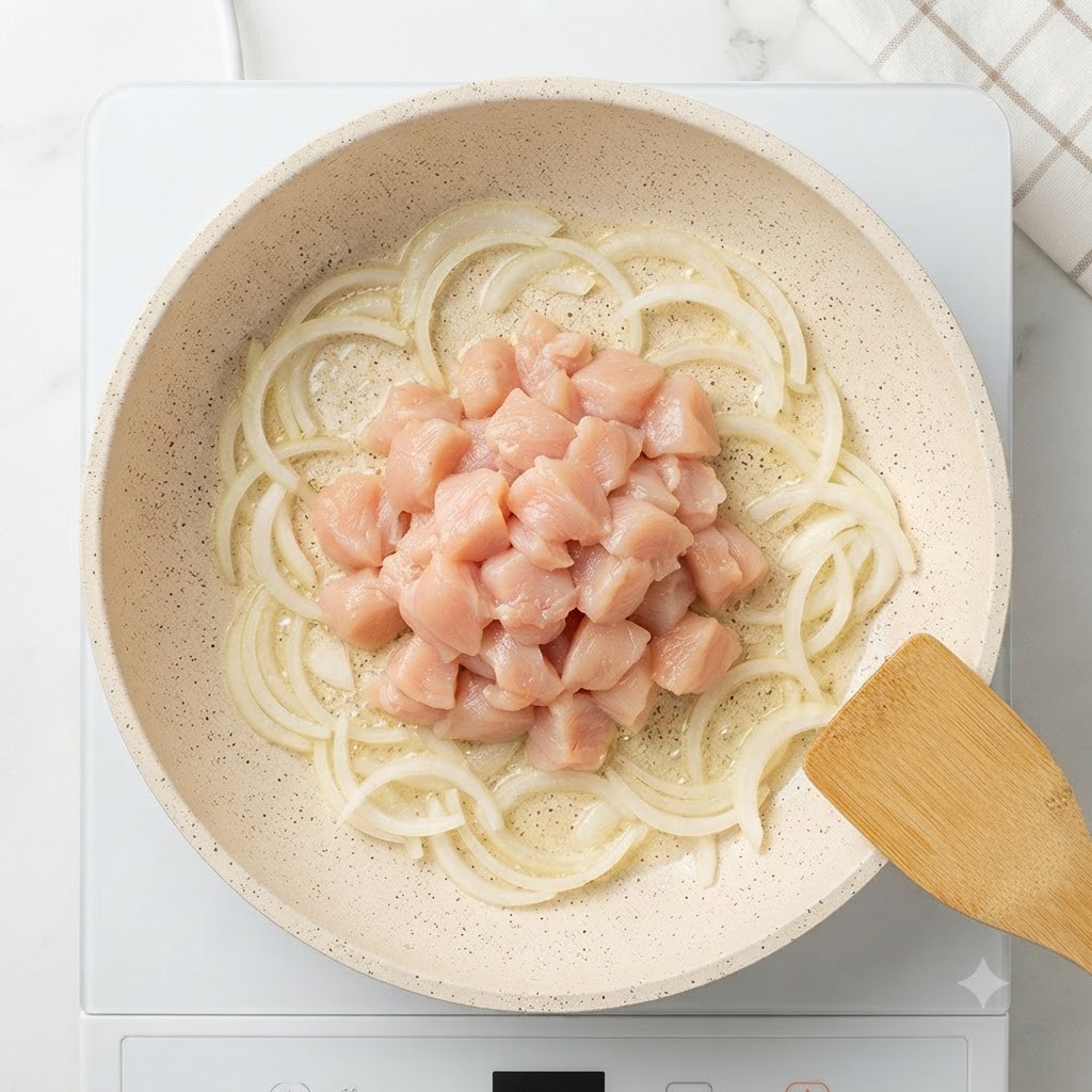 炒めた玉ねぎに囲まれて、フライパンの中心に生の鶏胸肉のサイコロ状の塊が置かれている様子。