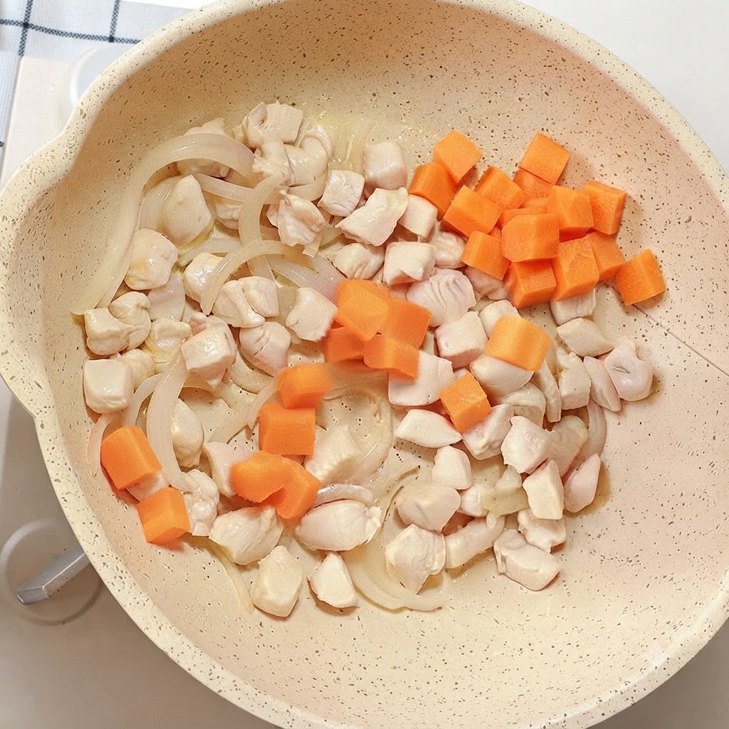 鶏肉と玉ねぎが入ったフライパンに、鮮やかなオレンジ色のニンジンのサイコロが加えられている様子。