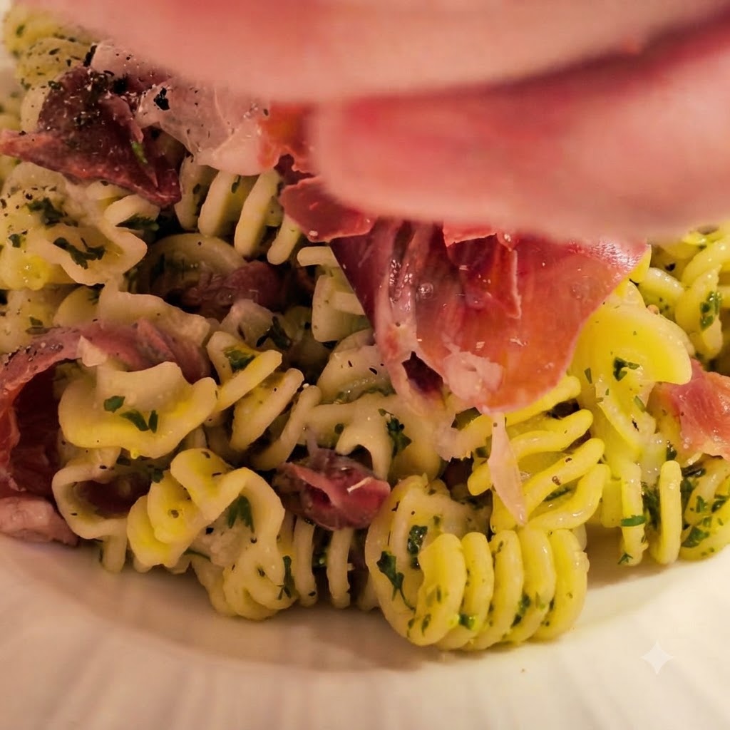 茹でたてのスパイラルパスタが、用意したバターとガーリックのソースが入ったボウルに投入され、新鮮なハーブやハムが添えられているところ。