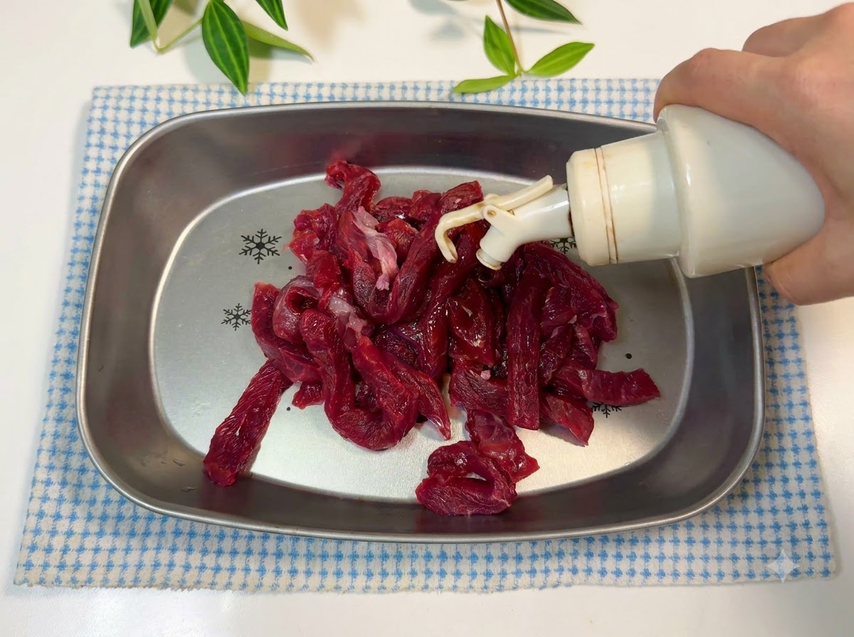 金属製のトレイに入った牛肉の細切りに油と醤油で味付けをしている様子。