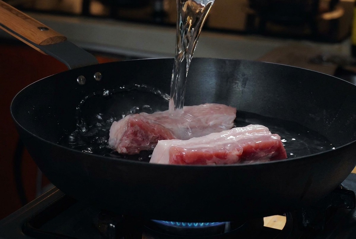 冷たい黒いフライパンに並べた生の高みのある豚バラ肉に、計量カップから水を注いでいる様子。