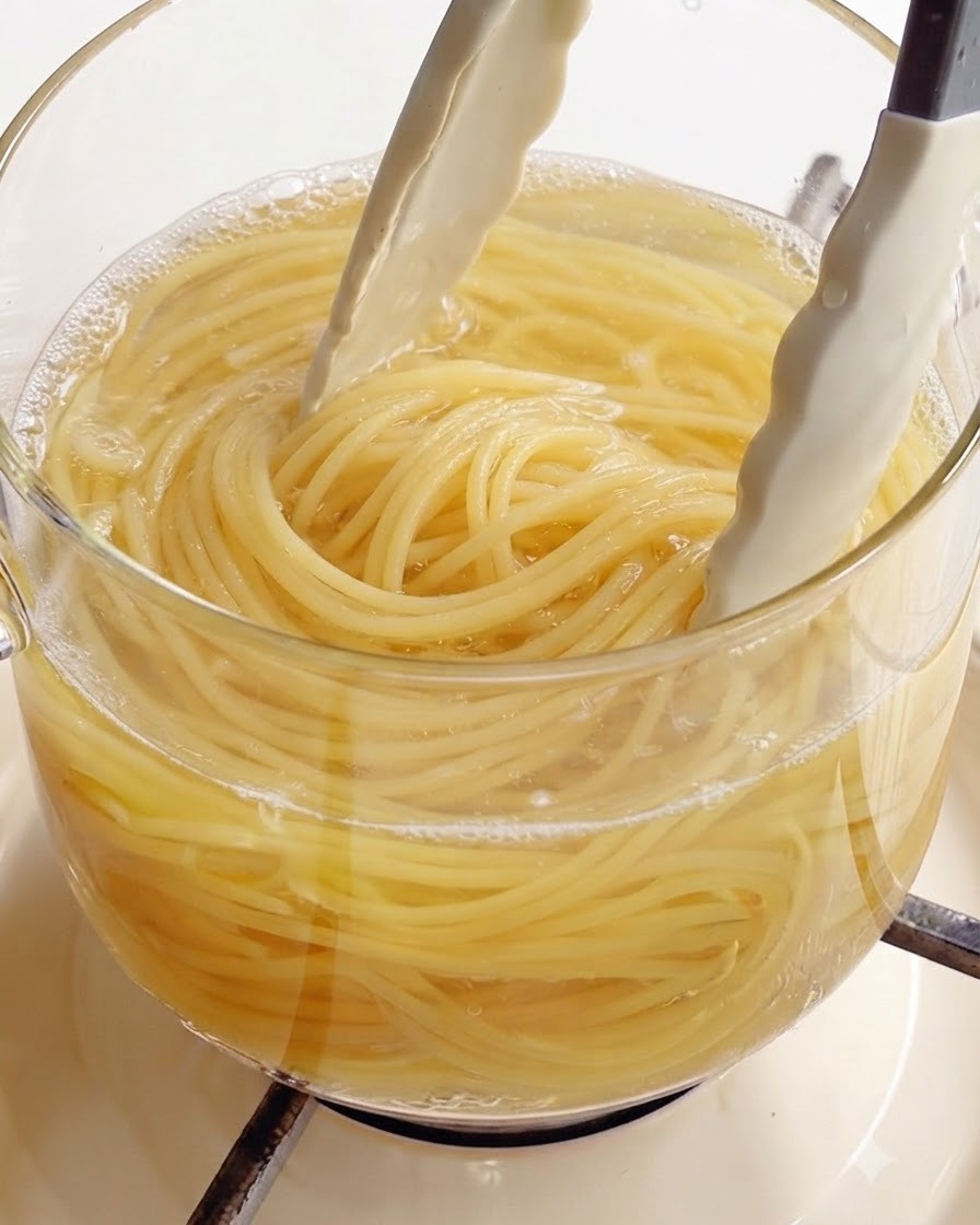 沸騰したガラス鍋から、白いキッチン用トングでアルデンテに茹で上がったスパゲッティを持ち上げているところ。