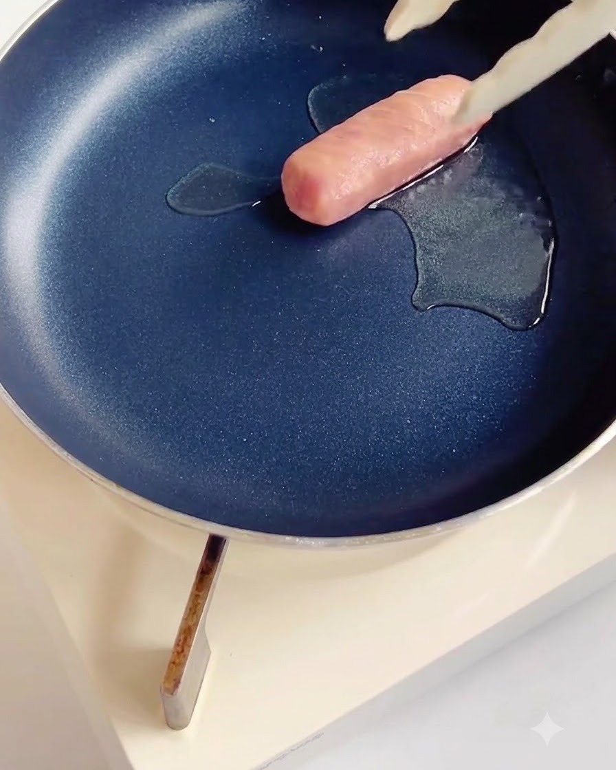 少量の油をひいた黒いノンスティックパンで、切れ目を入れたソーセージを炒めているところ。