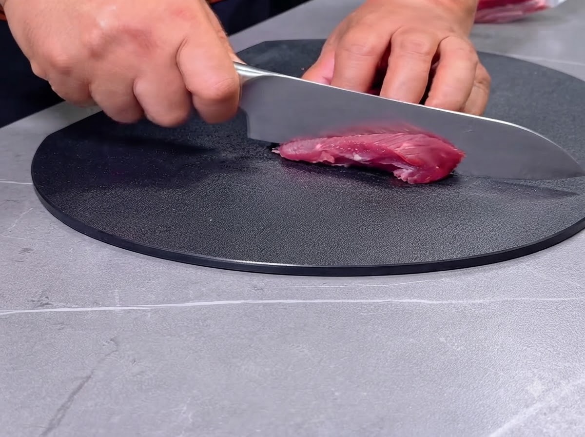 黒いまな板の上で、包丁を使って生の赤身肉を一口大に切り分けている人物の手元。