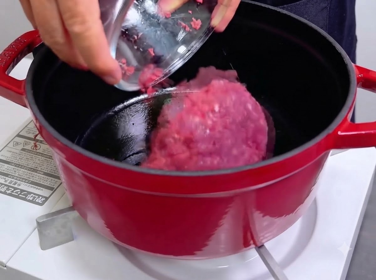 赤い鋳物ホーローの熱したダッチオーブンに、ボウルからピンク色の生のひき肉を投入しているところ。