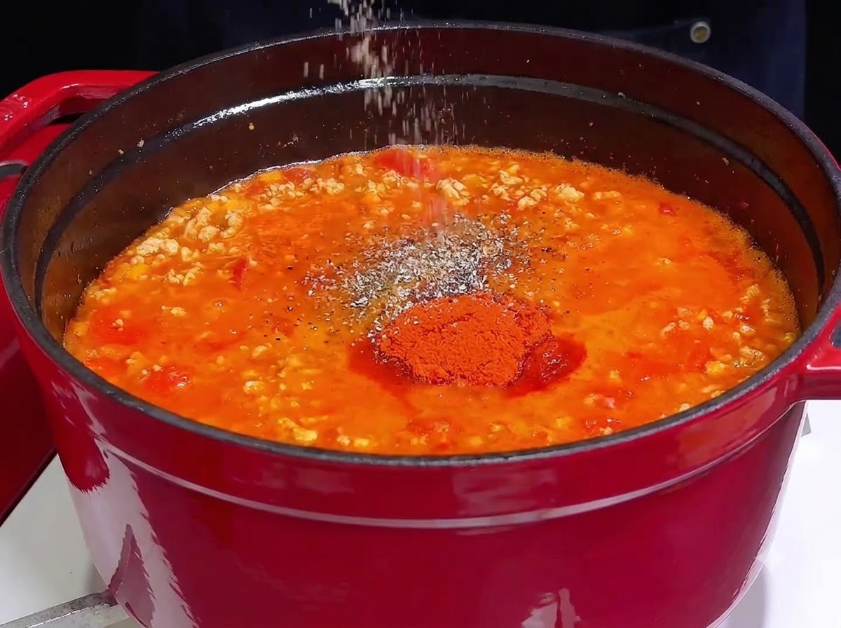 鋳物ホーロー鍋の中の鮮やかな赤い煮込みソースに、手が塩やスパイスを振りかけているところ。
