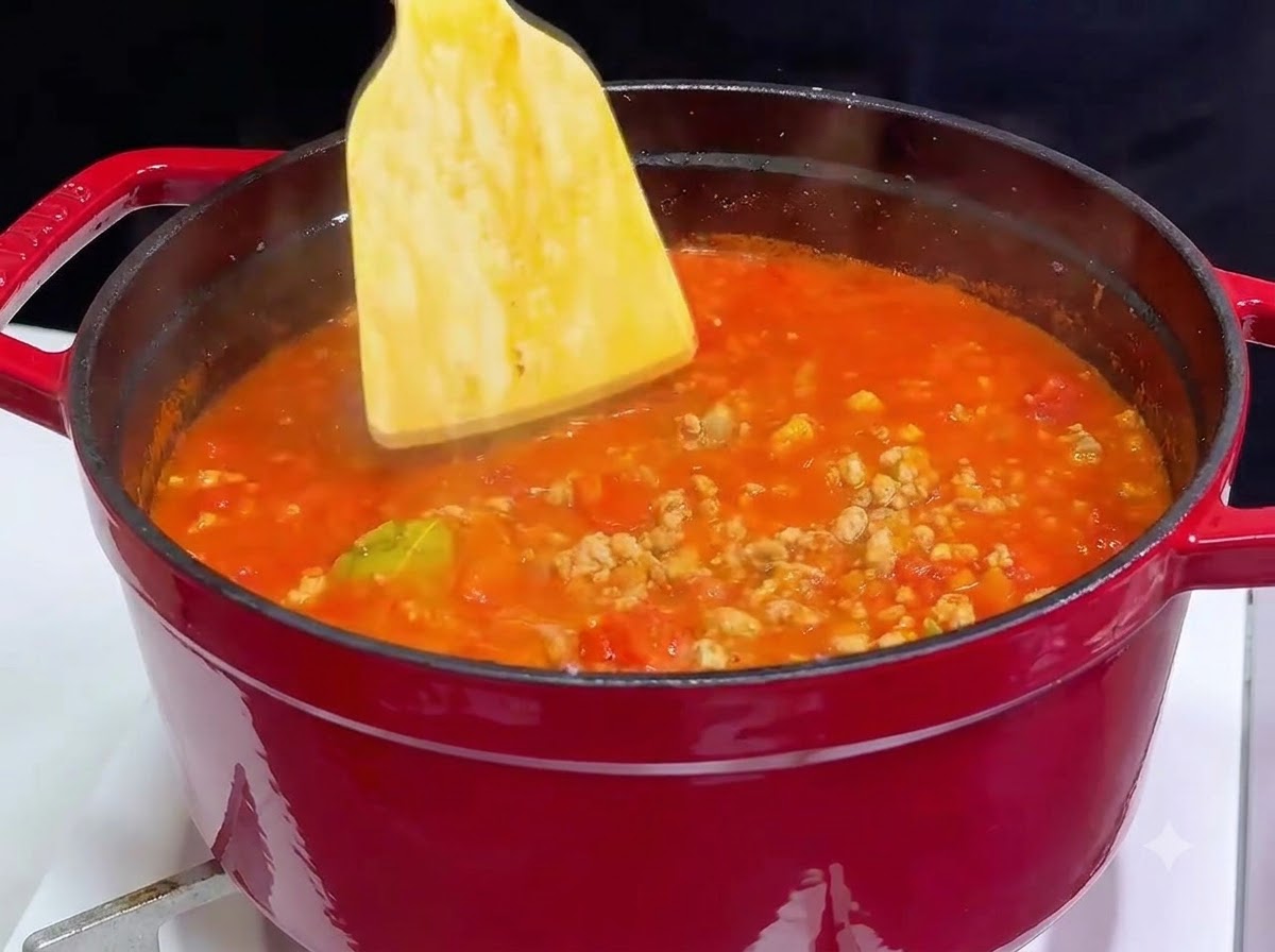 グツグツと煮えるトマトミートソースが入った赤い鋳物ホーロー鍋を、木べらでかき混ぜている様子。
