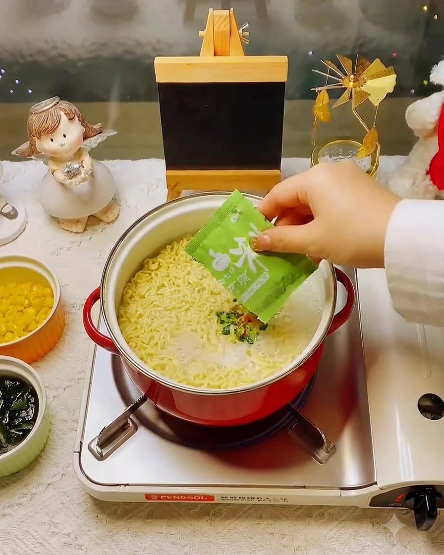 麺とスープの入った鍋に、緑色の小袋に入ったかやくを加えている様子。