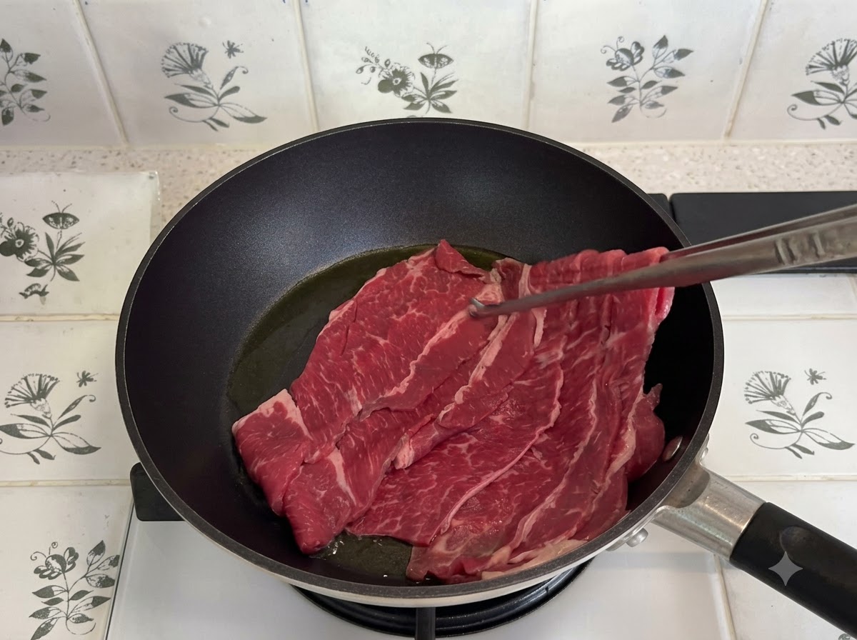 黒いフライパンの中で、トングを使って薄切りの牛バラ肉を炒めている様子。