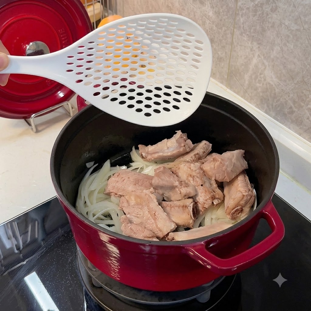 白いスキマー（網じゃくし）を使って、炒めた玉ねぎのベッドの上に下茹でした豚スペアリブを赤い鍋に入れているところ。