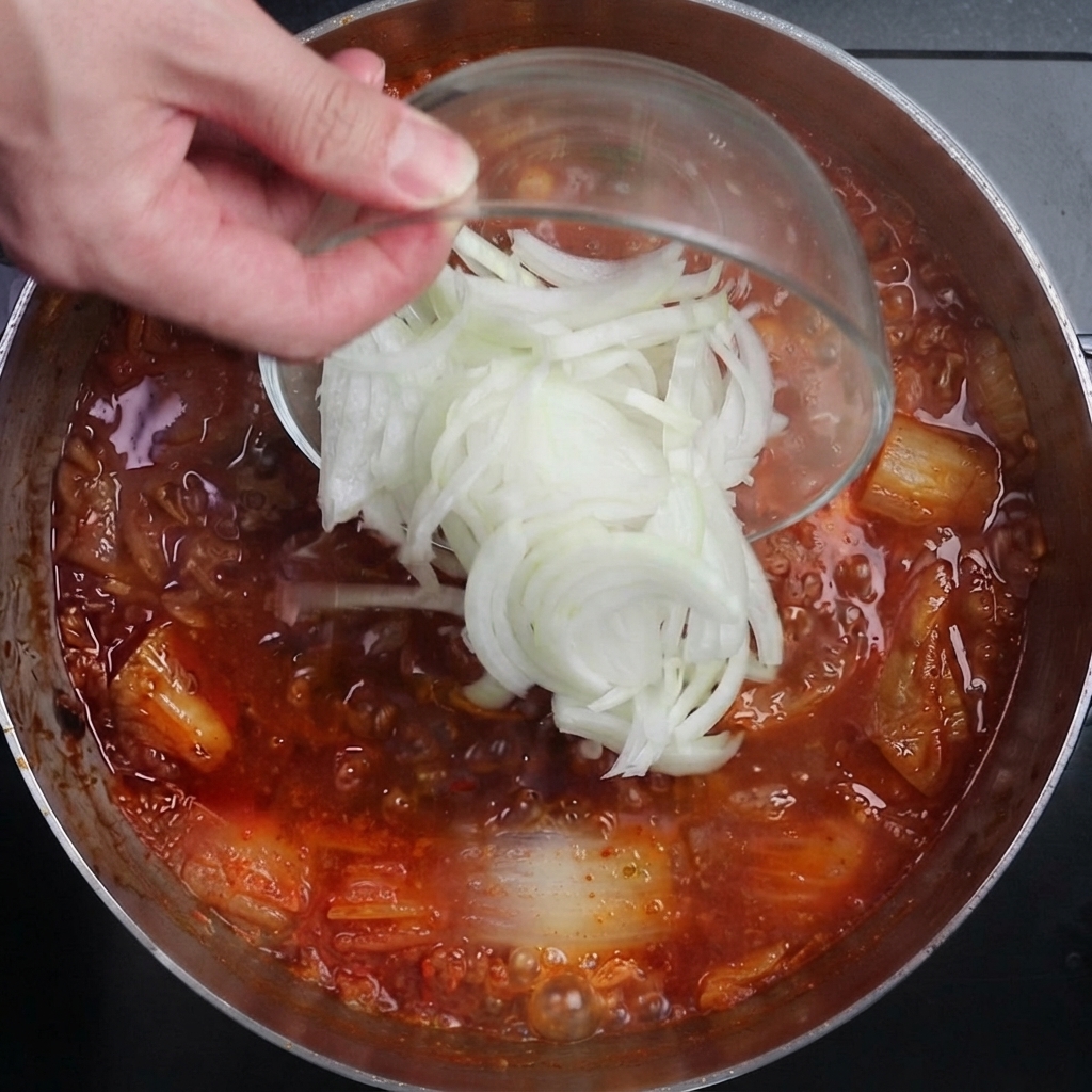 グツグツと煮立つスパイシーな赤いキムチスープに、薄切りにした玉ねぎを入れている様子。