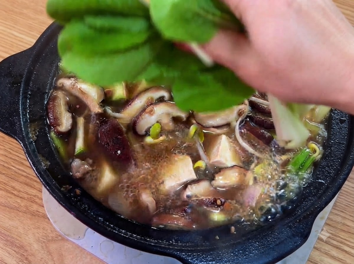 煮立っているスープの上に、新鮮な緑の葉野菜とスライスした青唐辛子を乗せている様子。