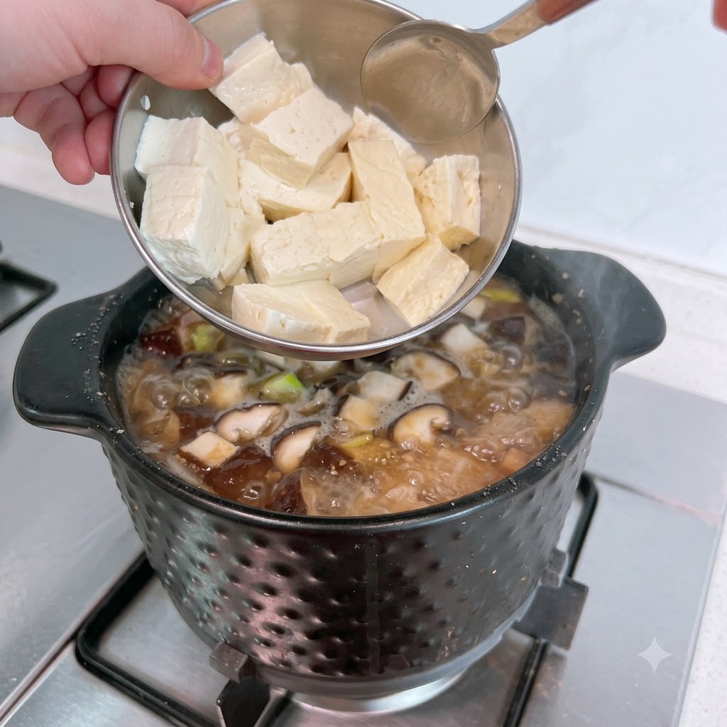 金属製のボウルから、ぐつぐつと煮えているチゲに白いスライス豆腐を加えている様子。