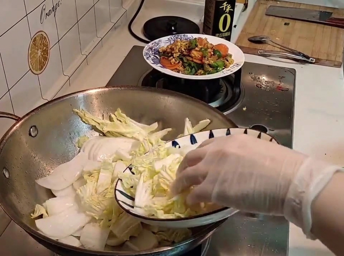 柄のあるお皿から、切り分けた生の白菜を中華鍋へ手袋をした手で移しているところ。