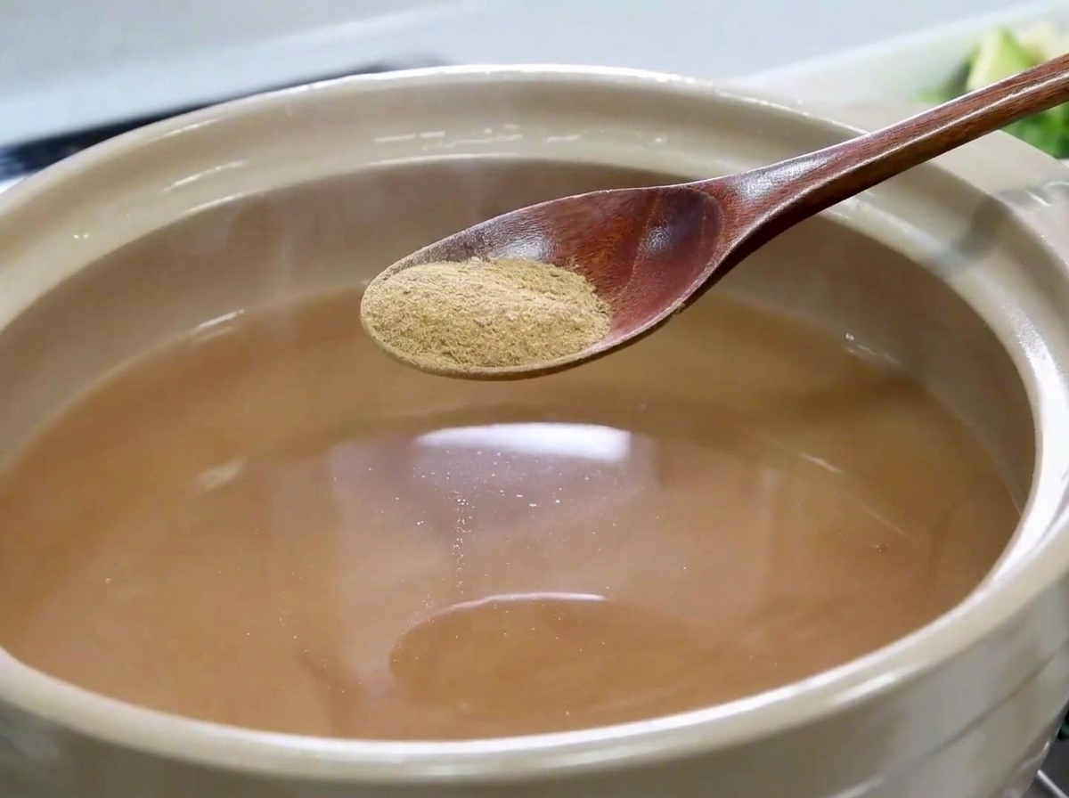 薄茶色のスープで満たされた鍋の上に、細かい煮干しパウダーを乗せた木べらがある様子。