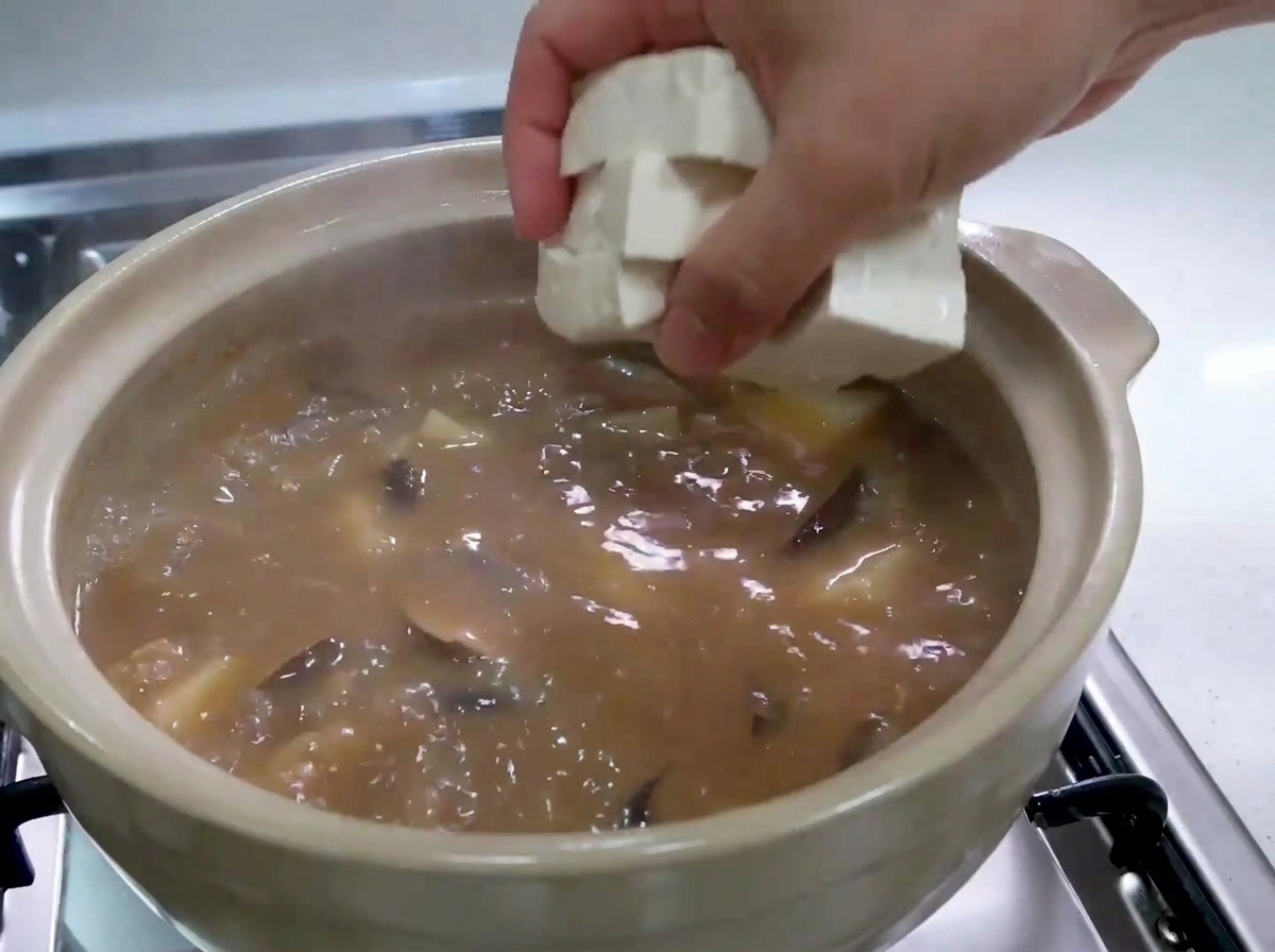 グツグツと煮立つ茶色のチゲの中に、新鮮な白い豆腐のキューブを手でそっと入れている様子。