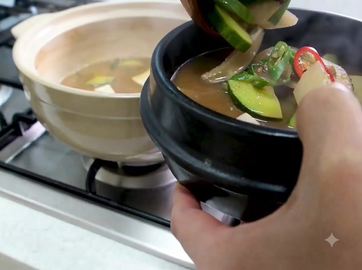 ズッキーニや野菜の入った熱々のテンジャンチゲを、伝統的な黒いサービングボウルにお玉ですすっている様子。