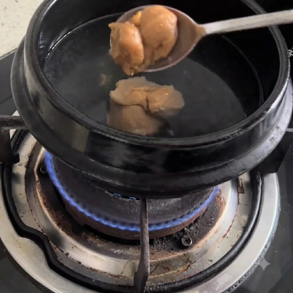 ガスコンロ上の沸騰したお湯が入った黒い鍋に、スプーン一杯の味噌が入れられている様子。