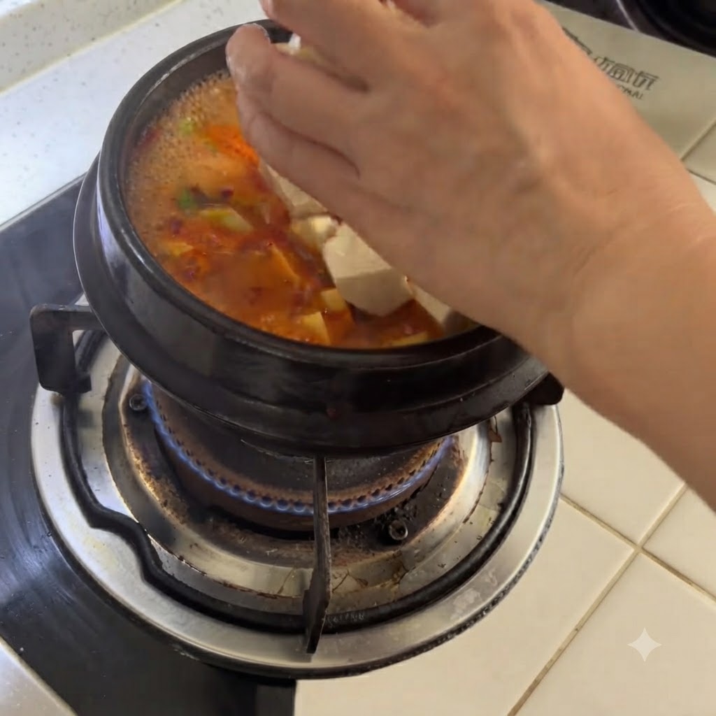 スパイシーなスープが入ったグツグツ煮える土鍋の中に、新しく切った白い豆腐の塊を手で落とし入れている様子。