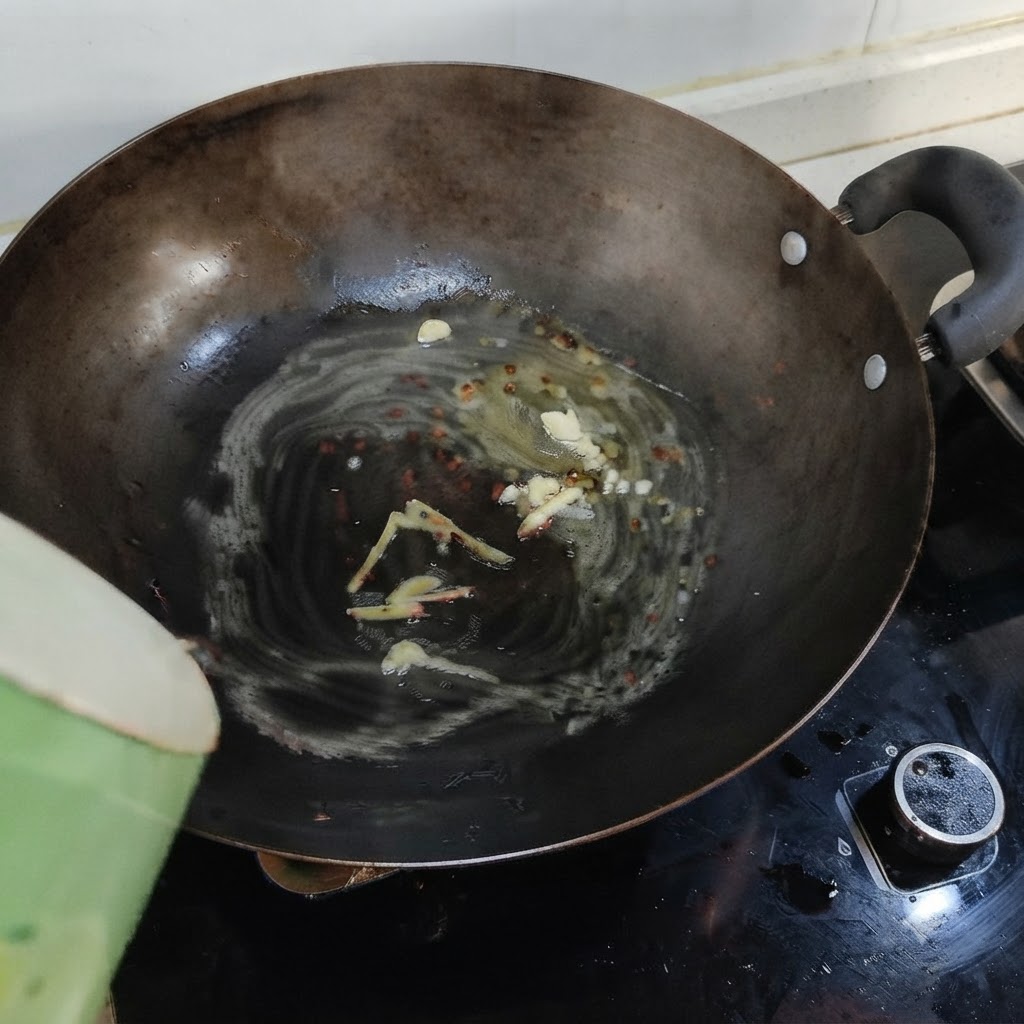 緑色の容器から、ラードや香味野菜の入った中華鍋へ熱湯を注いでいる様子。