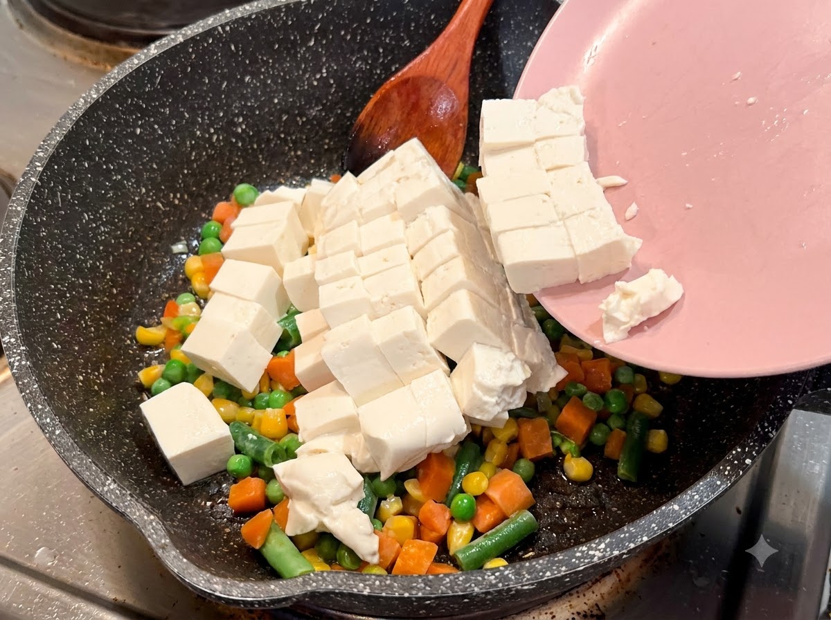 サイコロ状の絹ごし豆腐を、ピンク色の皿から野菜の入ったまだら模様のフライパンへ滑り込ませている様子。