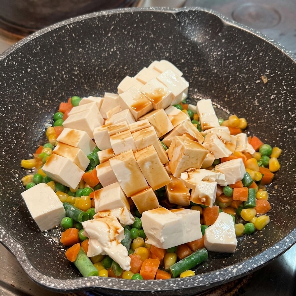 フライパンの中の絹ごし豆腐とミックスベジタブルの上に、しょうゆを回しかけている様子。