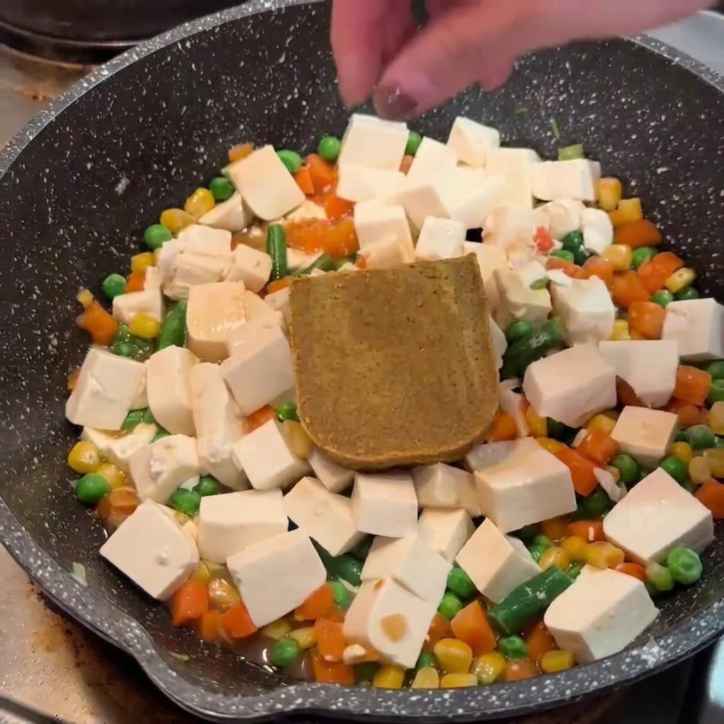 フライパンの中央にある豆腐と野菜の上に、固形の茶色いカレールーを置いている手。
