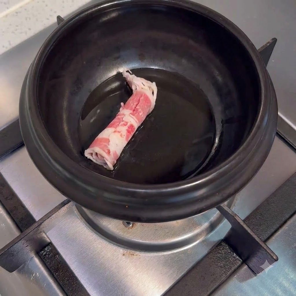 コンロの上の黒い陶器鍋に油をひき、生の牛薄切り肉を入れている様子。