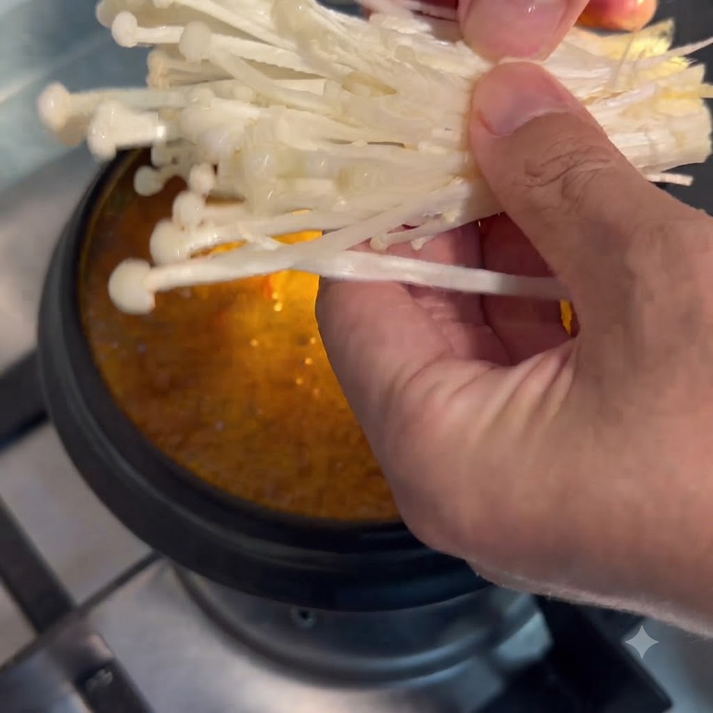 手で持ったえのき茸を、煮立っているチゲ鍋に入れている様子。