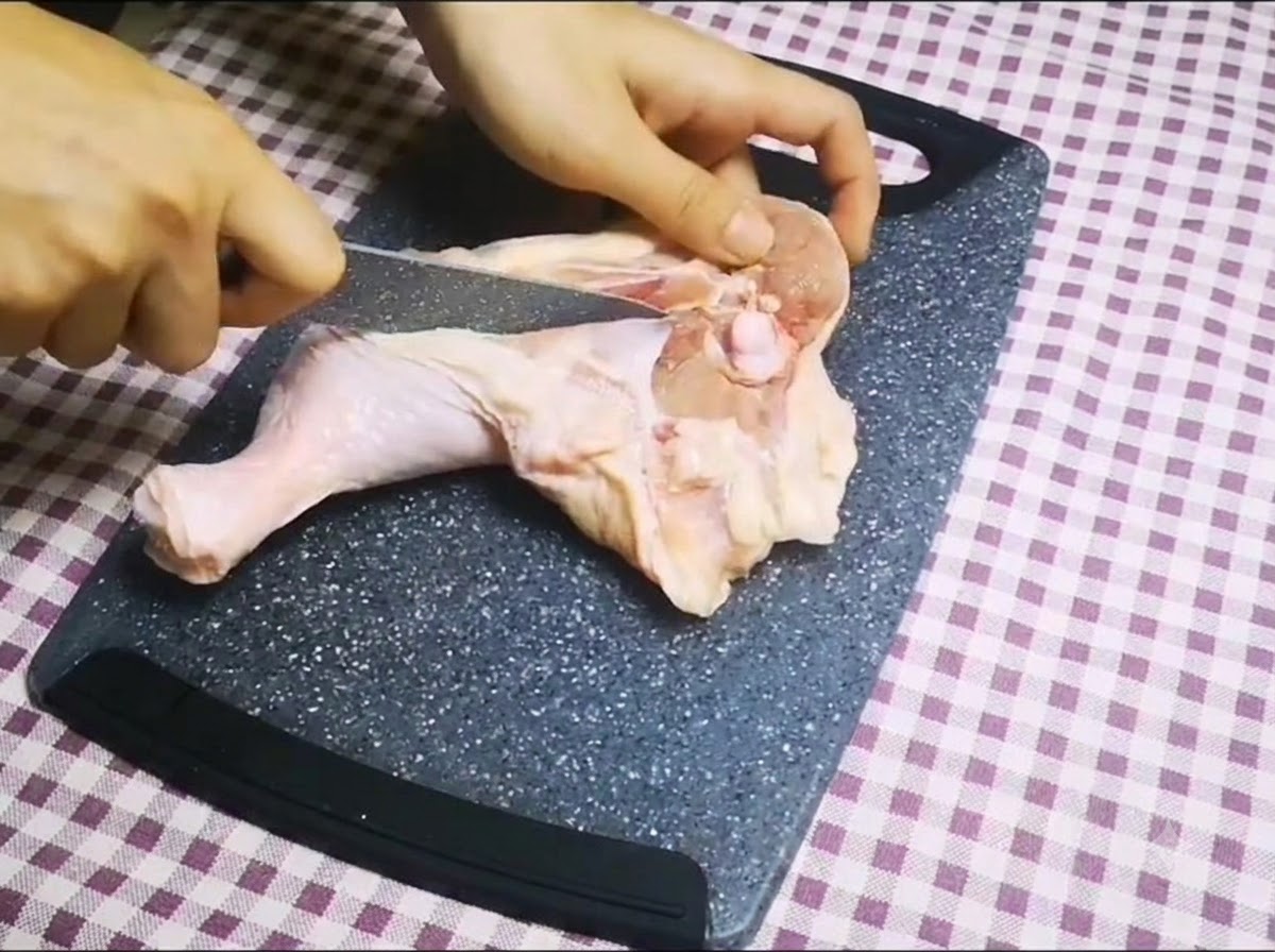 まな板の上で鋭いナイフを使って生鶏もも肉の骨に沿って切り込みを入れ、骨を取り除いている様子。