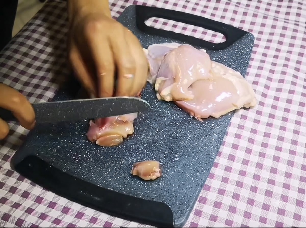 グレーのまな板の上でナイフを使い、骨なしの鶏肉を小さなチャンク状にスライスしている様子。