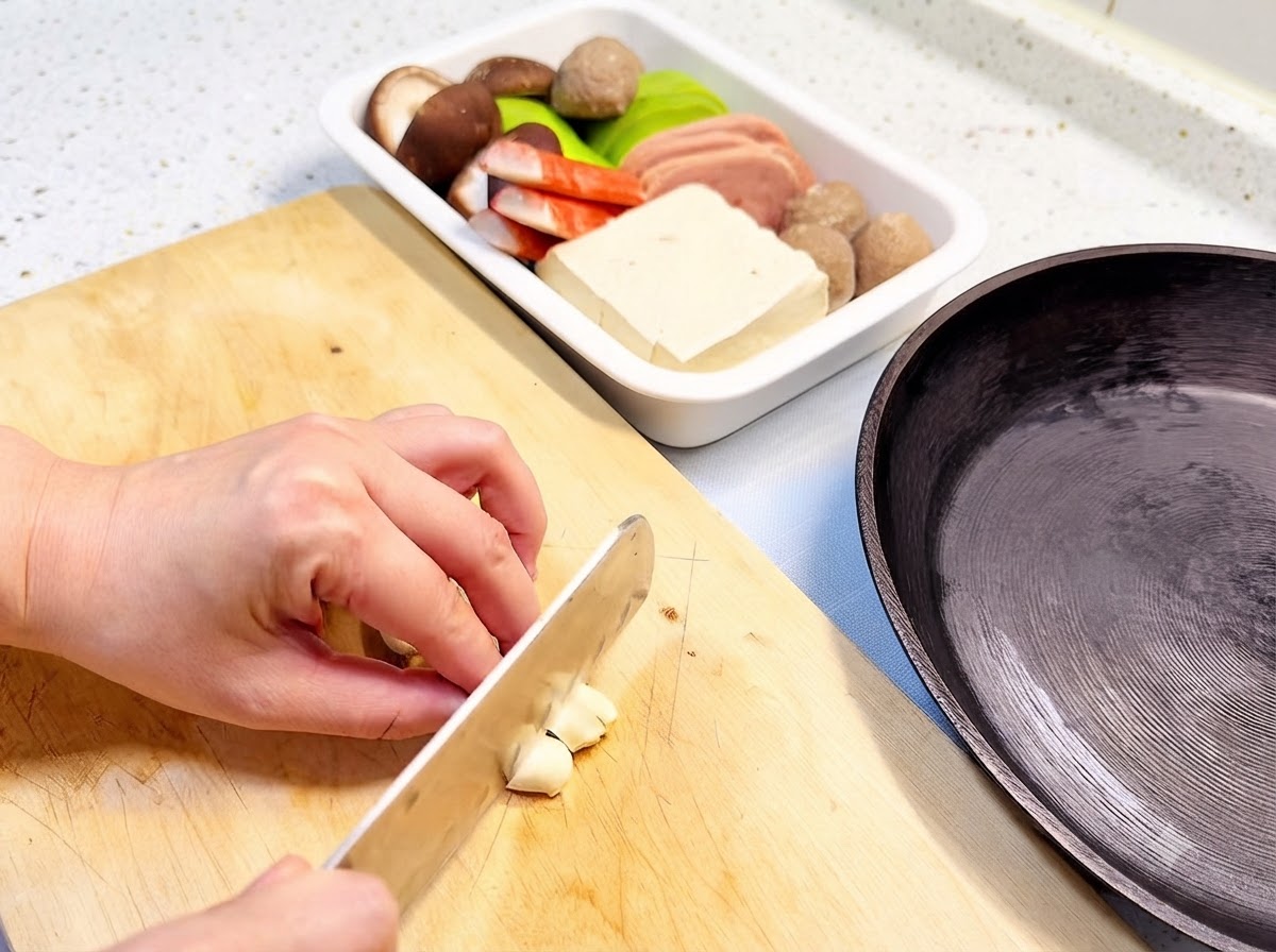 フライパンや具材トレイの隣にある木製のまな板の上で、にんにくのスライスをしている様子。