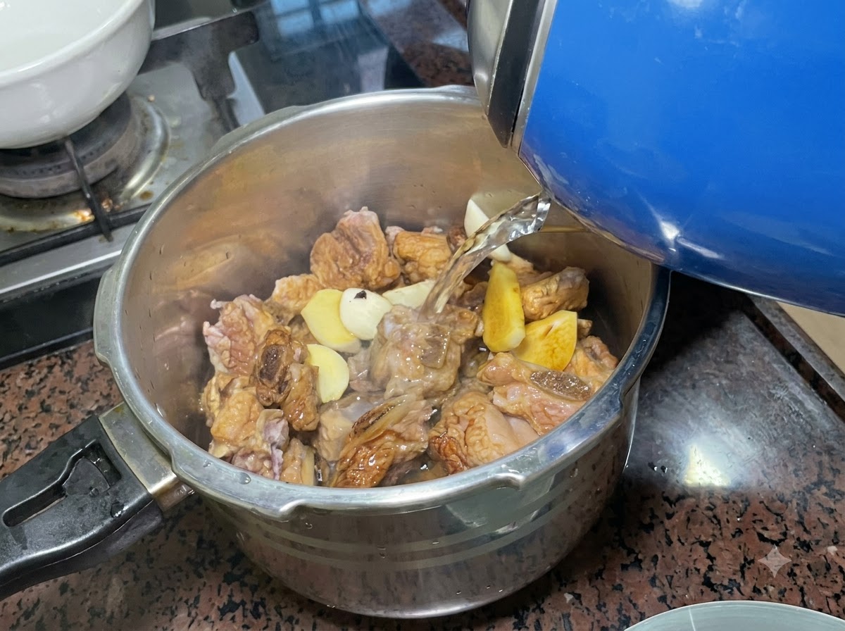 青い電気ケトルから、味付けされたリブと香味野菜で満たされたステンレス製の圧力鍋へ水が注がれている様子。