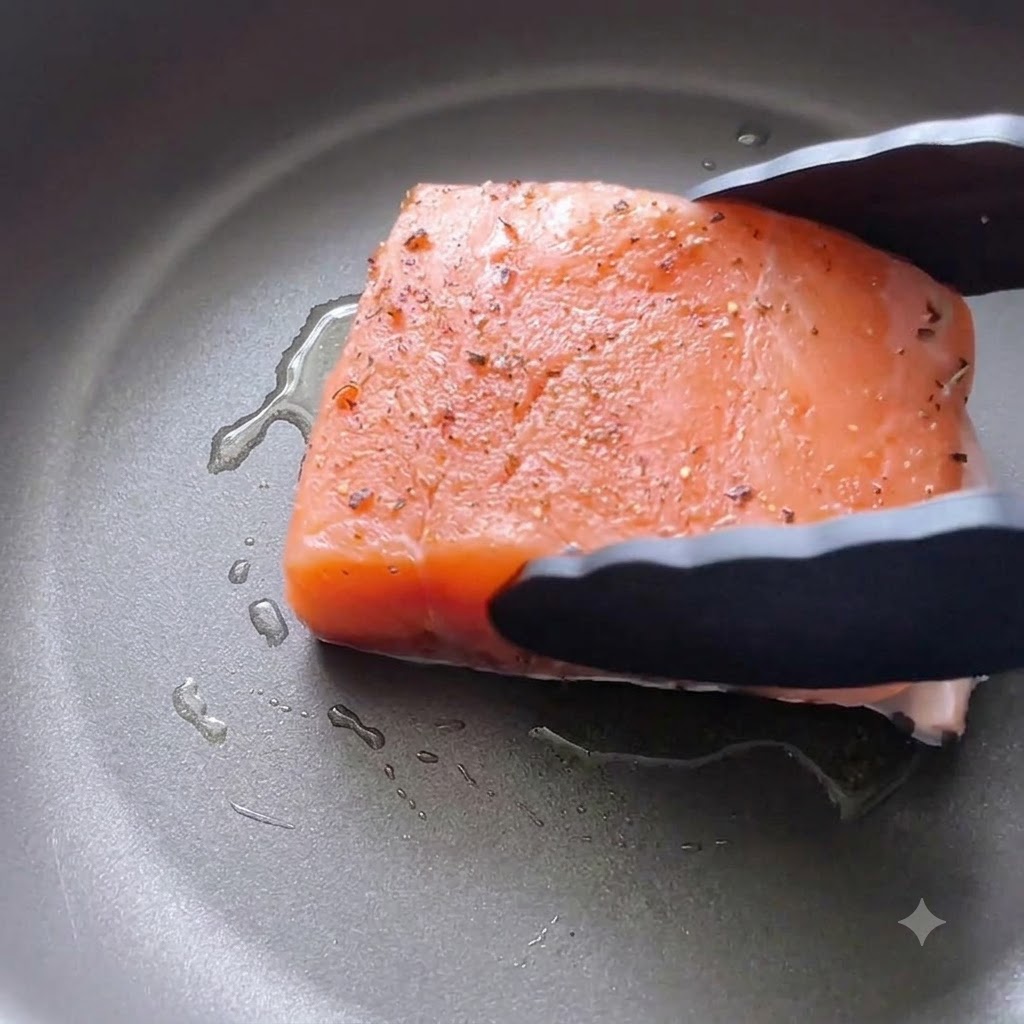 黒いキッチン用のトングを使って、下味をつけたサーモンフィレを油を引いたフライパンに皮を下にして降ろしているところ。
