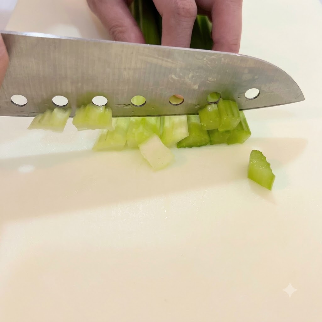 白いまな板の上で、大きな包丁を使って鮮やかな緑色のセロリの茎を小さく均等な大きさに刻んでいる手。