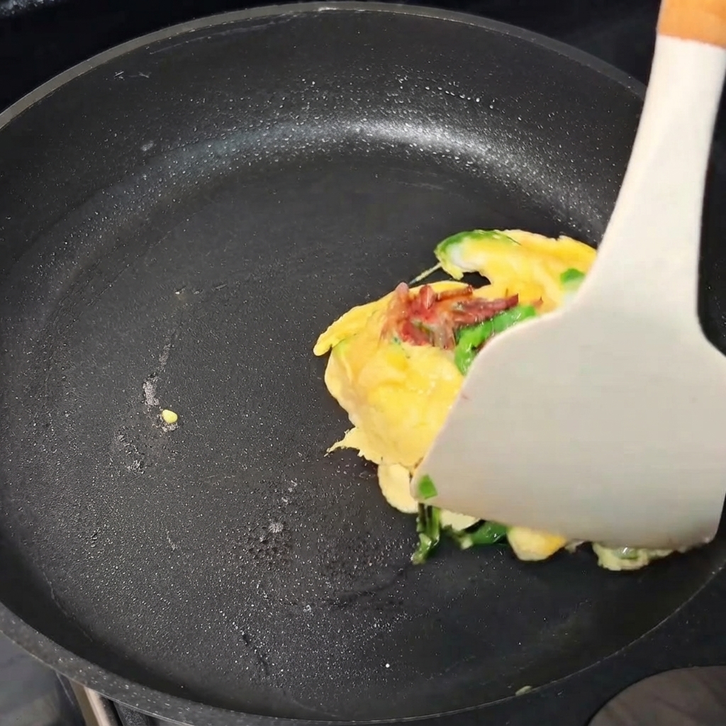 フライパンの中でヘラを使って卵のオムレツを包状に折りたたんでいる様子。