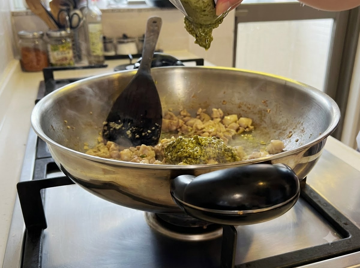 ガラス瓶に入ったグリーンペーストソースが、中華鍋の鶏肉とニンニクの混合物に混ぜ合わされている様子。