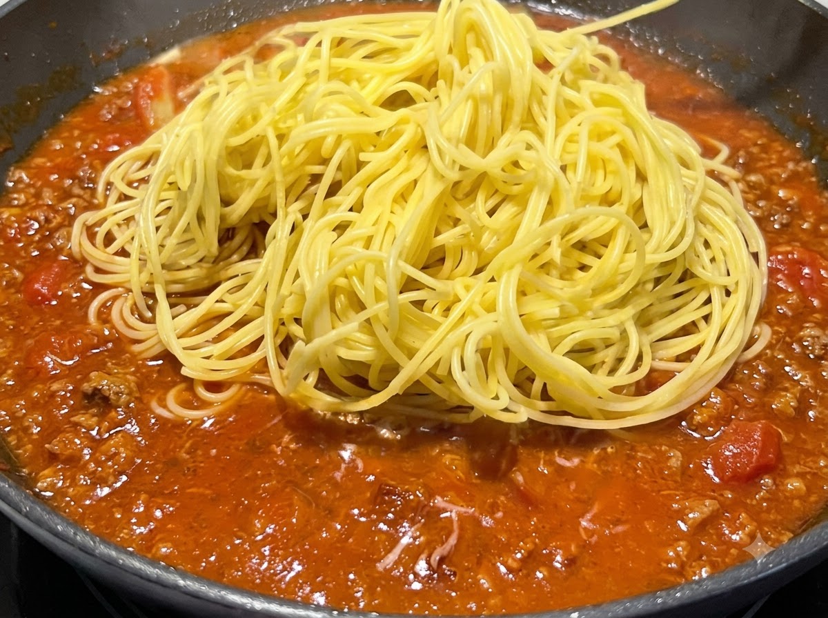 フライパンの中で煮立っている赤いソースの上に、茹で上がった黄色のスパゲッティの山が置かれている様子。