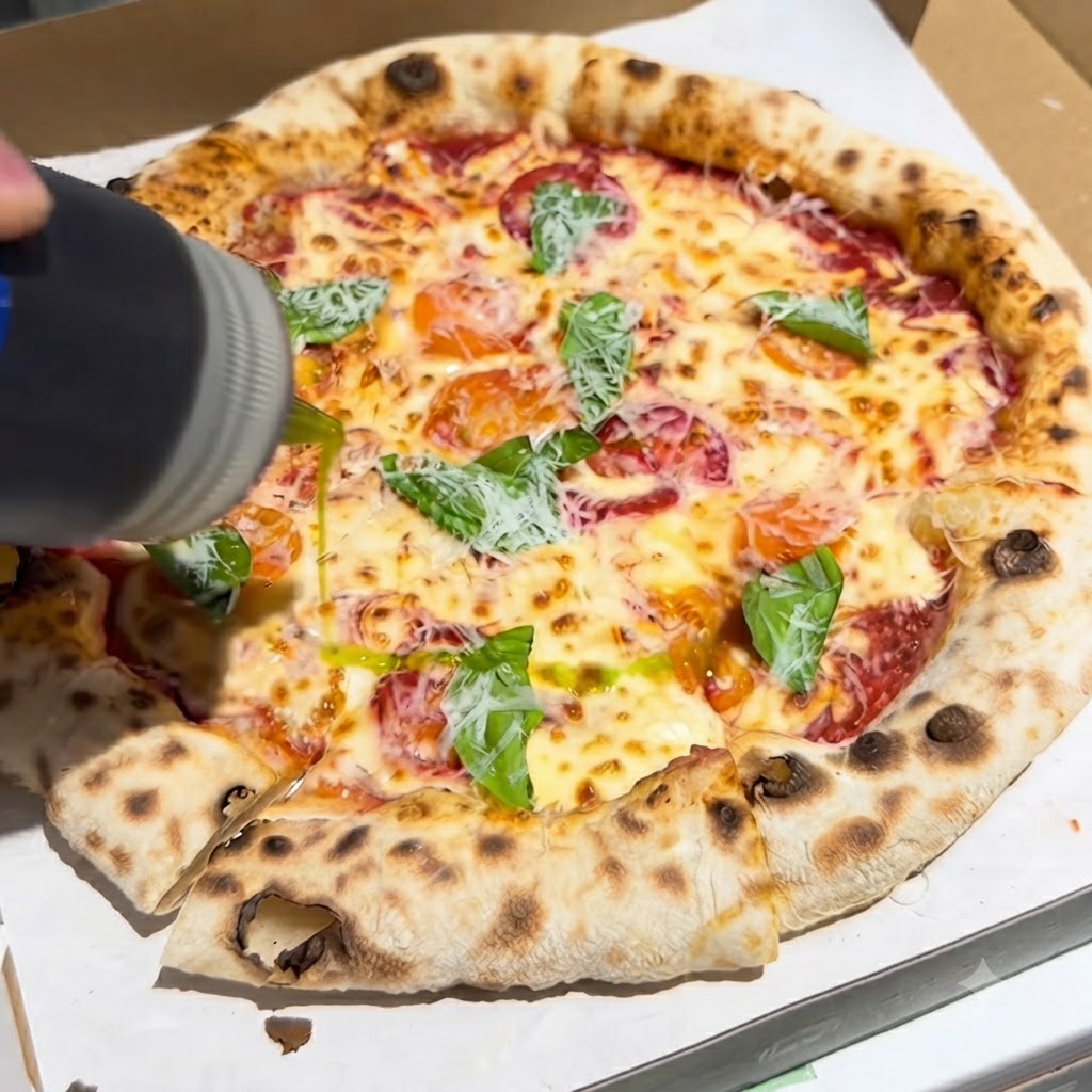 箱の中でバジルとチーズがトッピングされた焼き立てのピザに、ボトルからエキストラバージンオリーブオイルが注がれている様子。