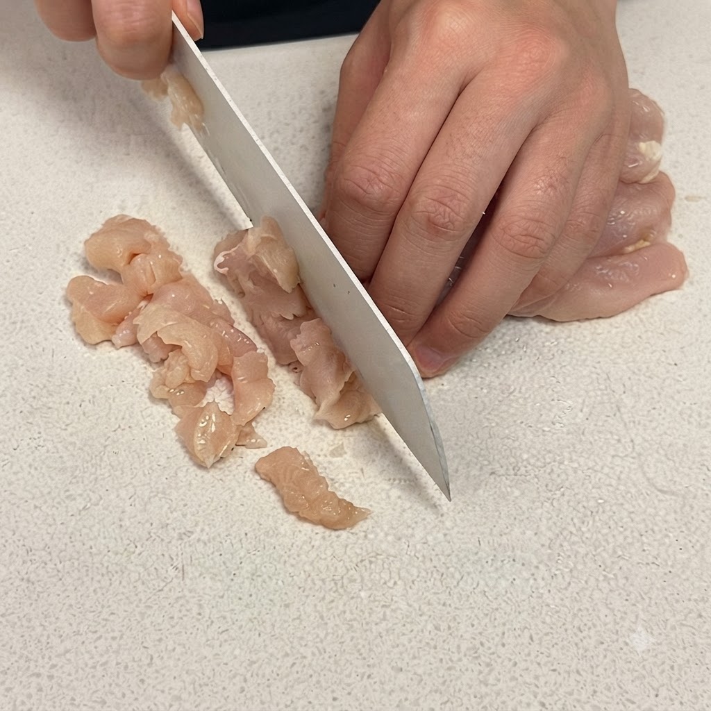 白いまな板の上で、よく切れるシェフナイフを使って鶏むね肉を小さなサイコロ状にスライスしているクローズアップ。