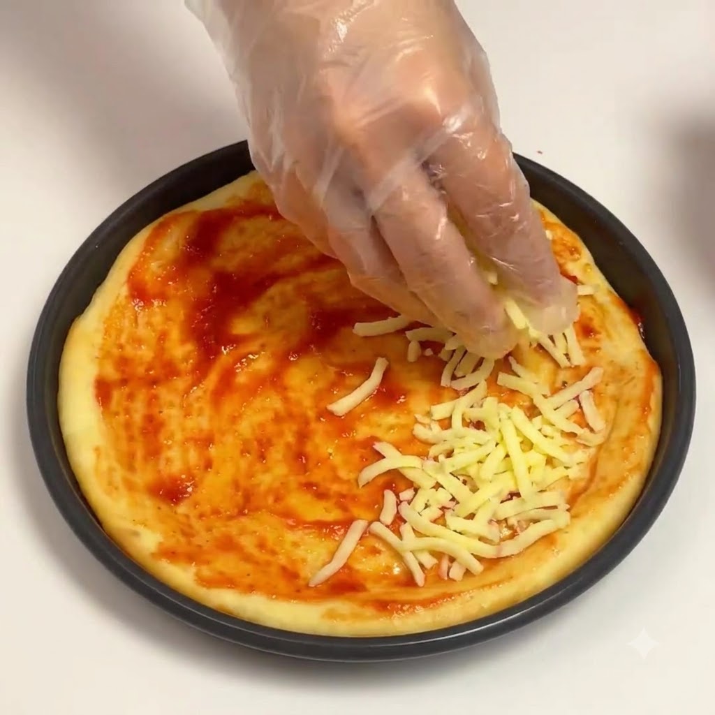 手袋を着用した手が、丸いピザパンの中のトマトソースのベースの上にシュレッドモッツァレラチーズをパラパラと振りかけている様子。