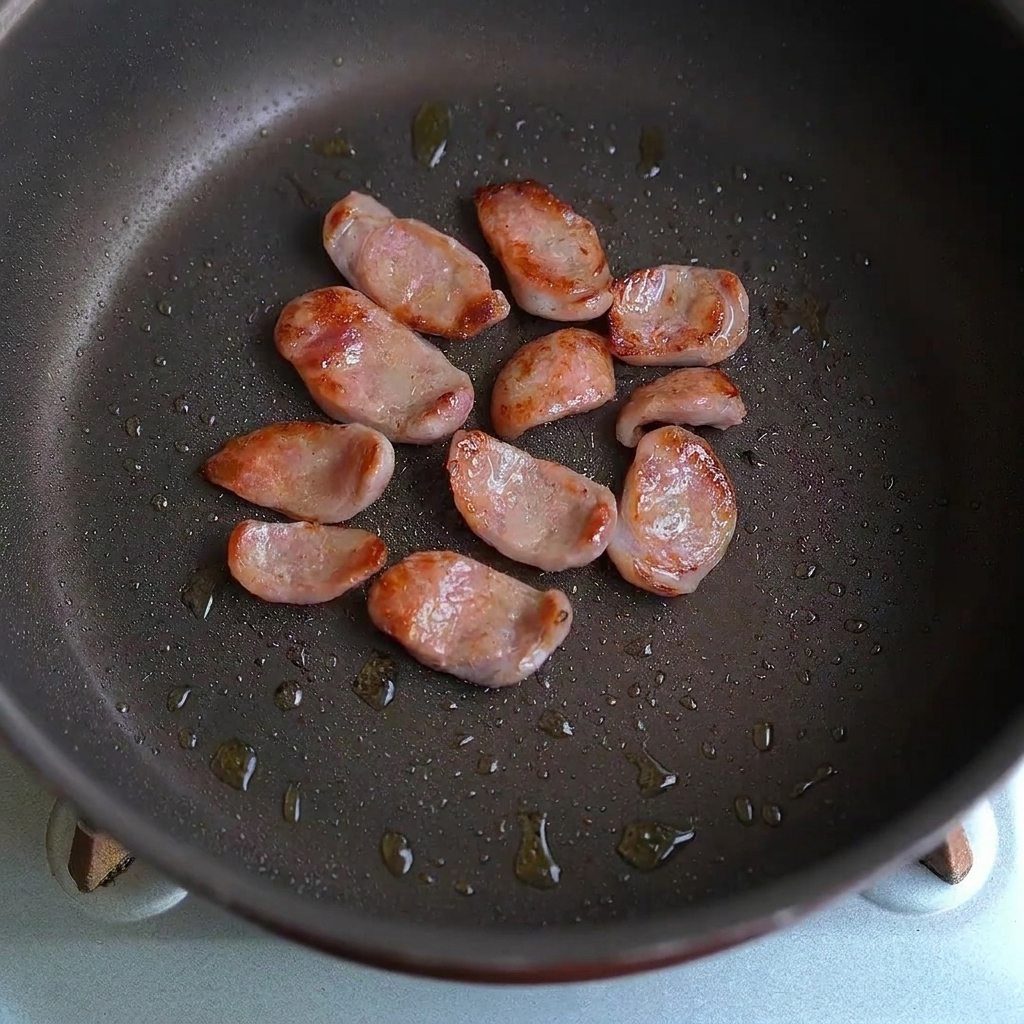 フライパンの中でソーセージのスライスとニンニクのかけらが一緒に炒められている様子。