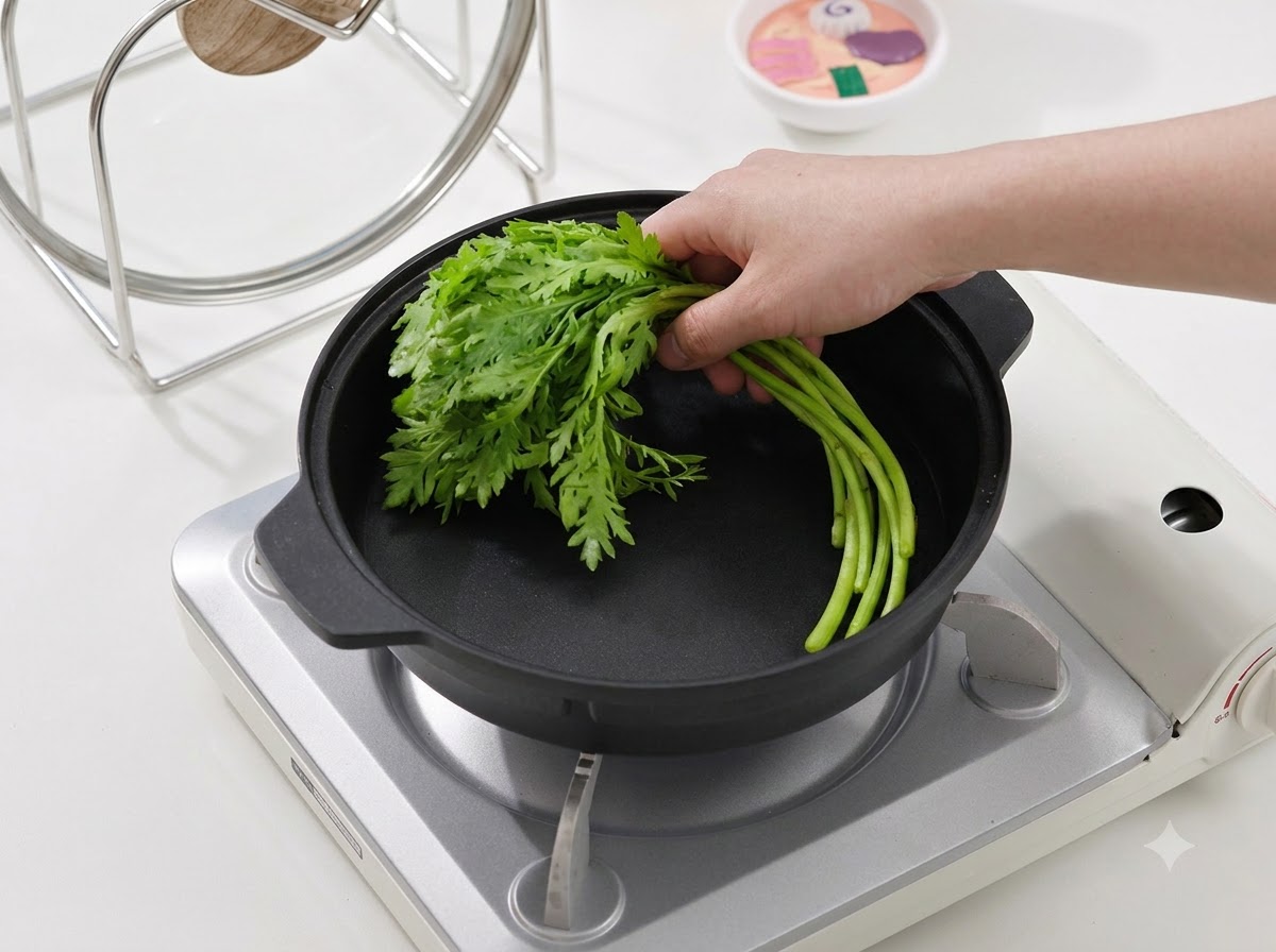 ポータブルコンロの上の黒い調理鍋に、一束の新鮮な緑色の葉物野菜を置いている手元。