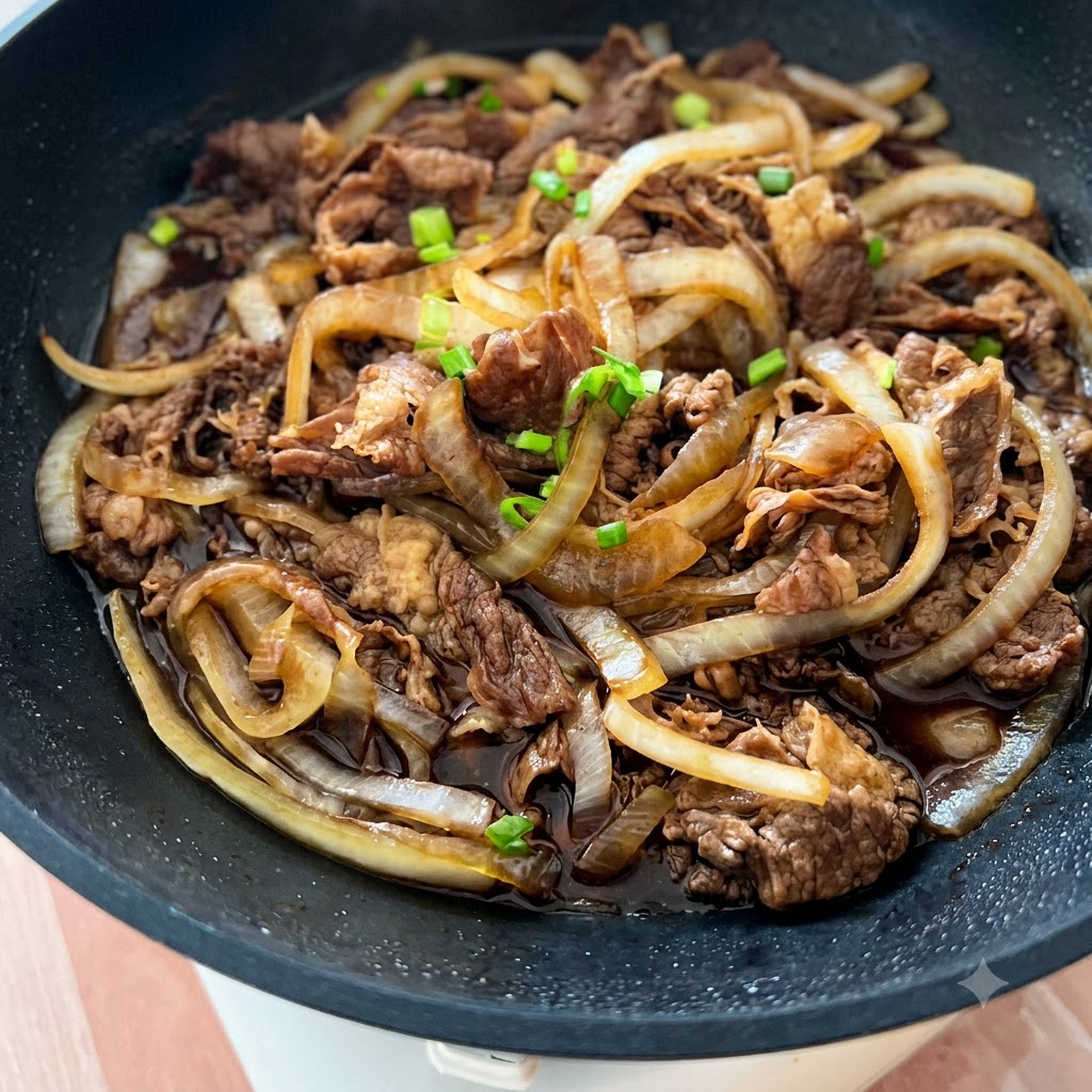 調理された牛肉と玉ねぎが入ったフライパンに、刻みネギと白いゴマが散らされている様子。