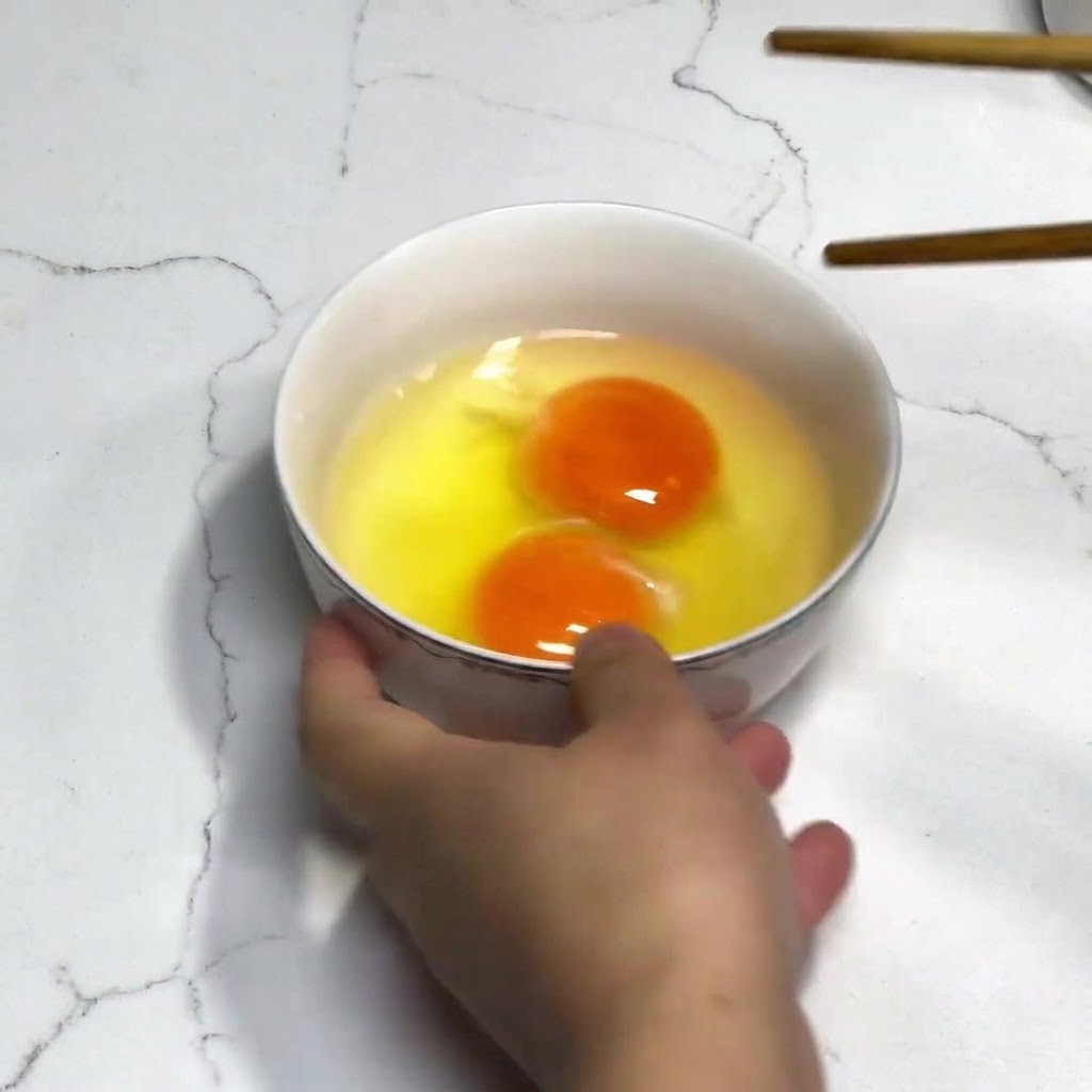 白いボウルに入った2個の新鮮な卵を、調味料なしで木製の箸を使って溶きほぐしているところ。