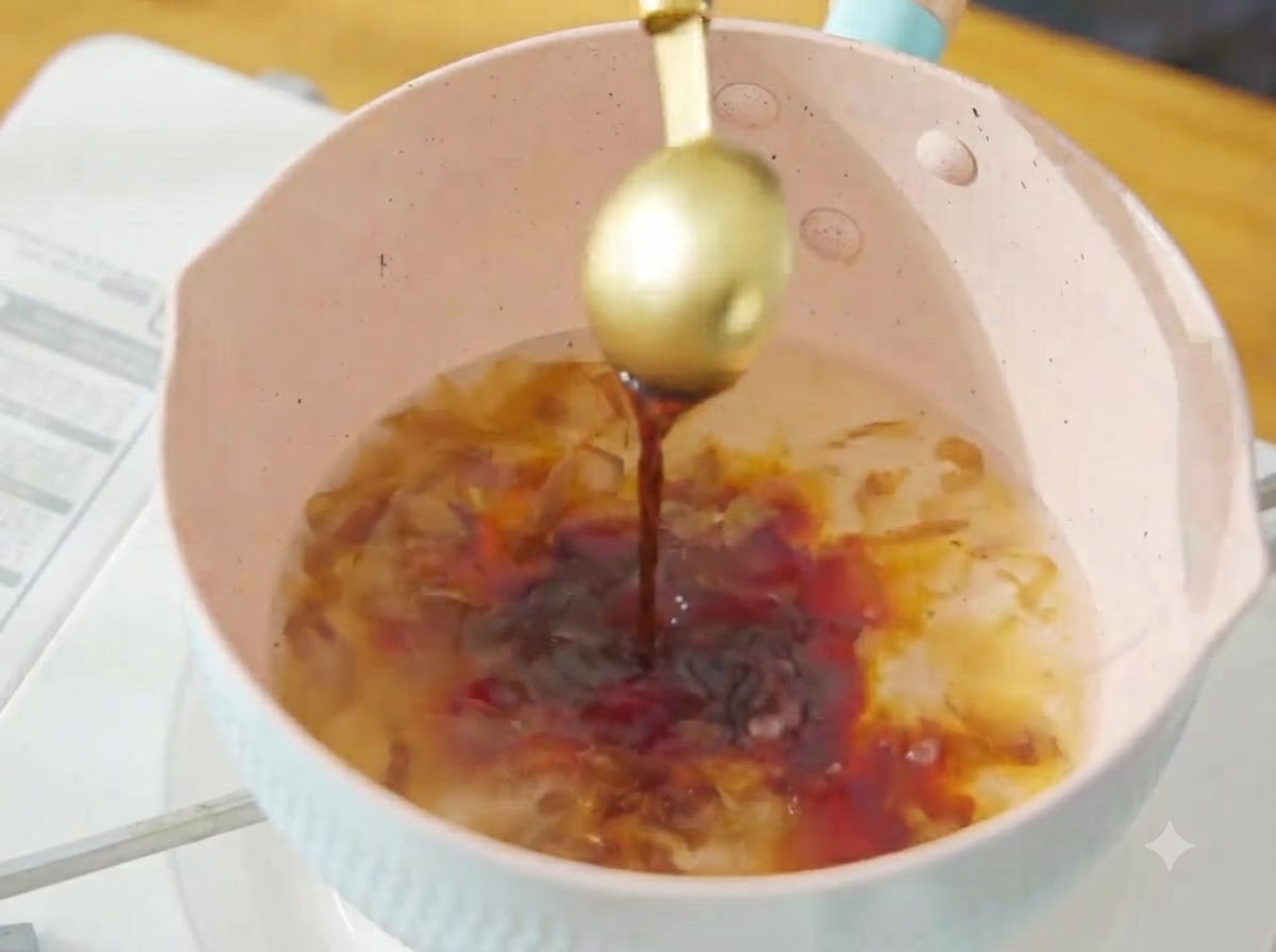 ピンク色の鍋の煮立っているかつお出汁に金色のスプーンで濃口醤油を注いでいる様子。
