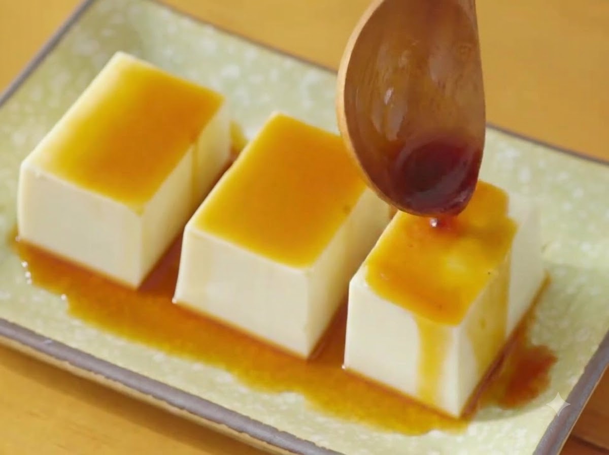 木製のスプーンを使って、緑色の皿に並べられた3列の絹ごし豆腐に醤油ベースのタレを回しかけているところ。