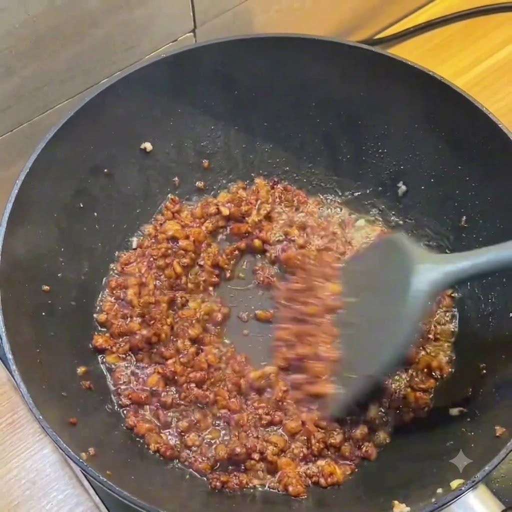 濃い茶色のひき肉ソースがグレーのスパチュラで混ぜられている暗い色の中華鍋の様子。