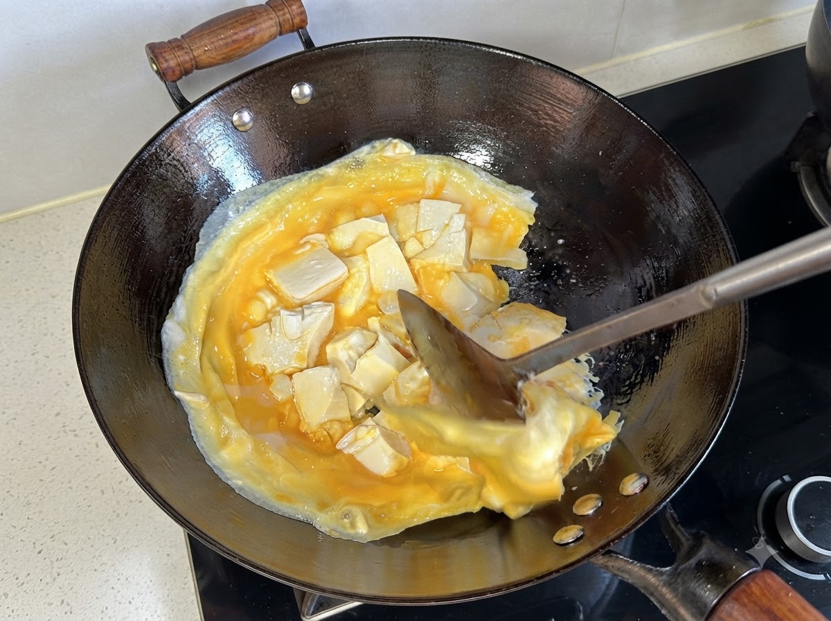 ヘラを使って、中華鍋の中で調理された卵と豆腐の層を持ち上げて裏返している様子。